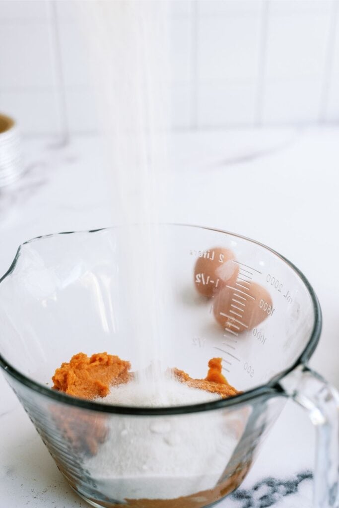 A measuring cup with pumpkin puree and sugar being poured in, with eggs in the background.