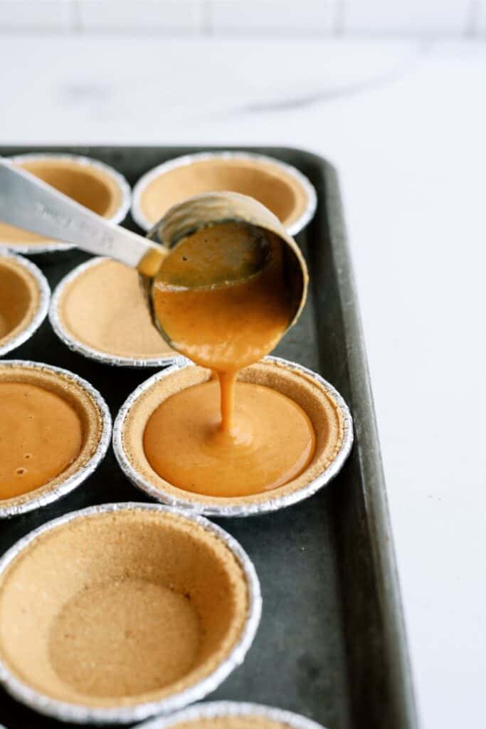 A spoon is pouring filling into individual pie crusts arranged on a baking tray.