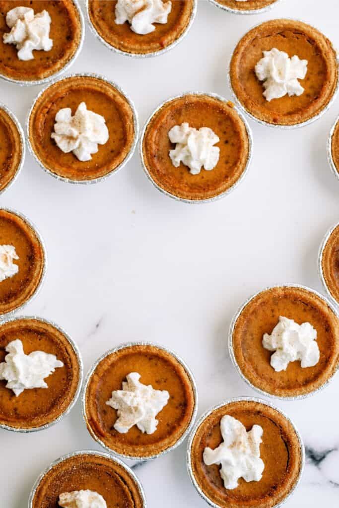 Several mini pumpkin pies with dollops of whipped cream on top are arranged in a circular pattern on a white surface.