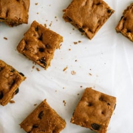 Several square-cut pieces of baked bar cookies lie on a white surface, with a few crumbs scattered around.