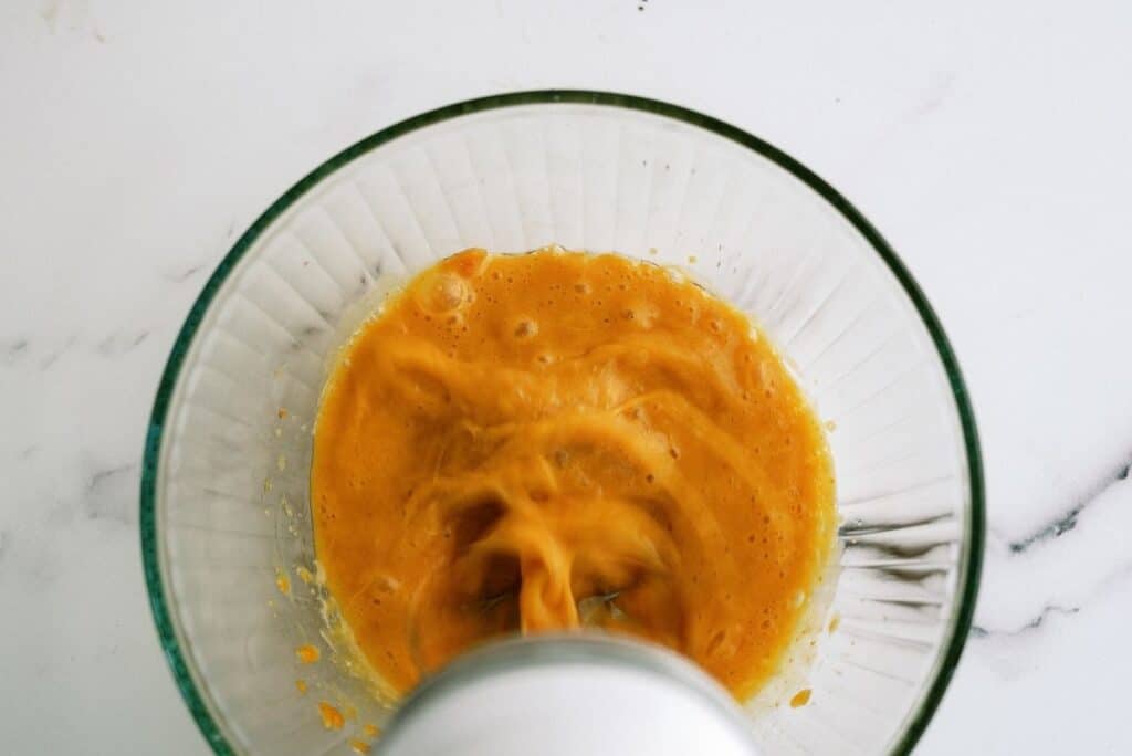 Top-down view of a hand mixer blending an orange mixture in a clear glass bowl.