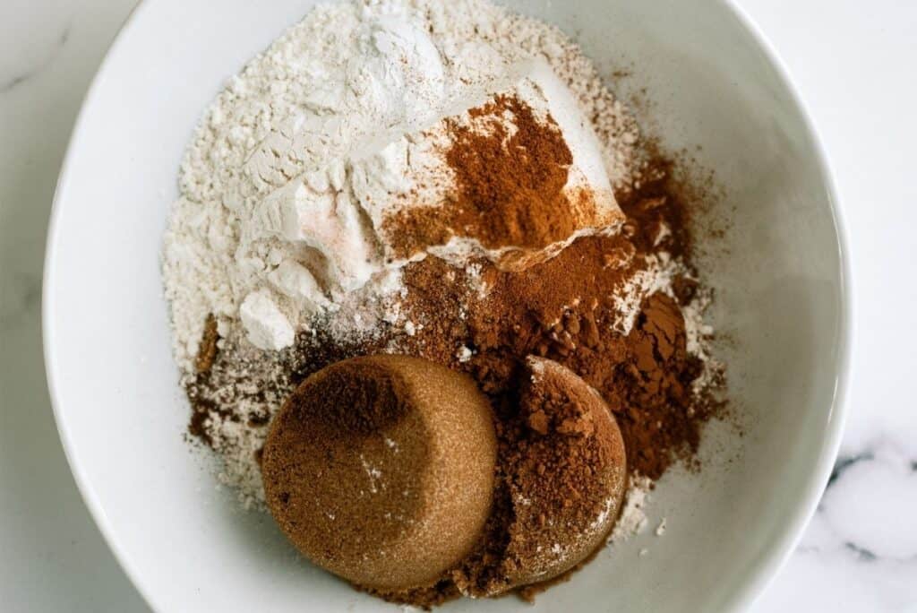 A white bowl containing various baking ingredients including flour, brown sugar, and ground spices.