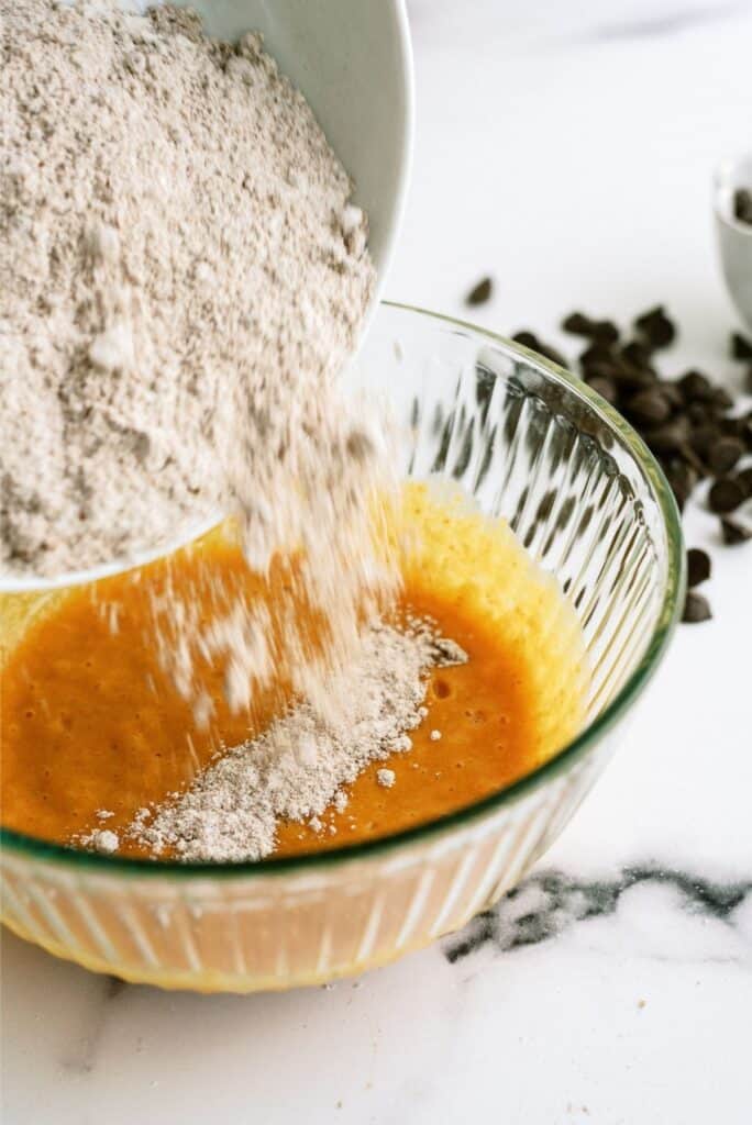 Dry ingredients being poured into a bowl of wet ingredients on a marble countertop, with chocolate chips in the background.