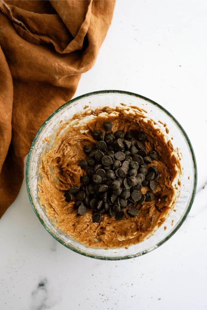 A glass bowl filled with cookie dough topped with chocolate chips, placed on a white surface next to a brown cloth.