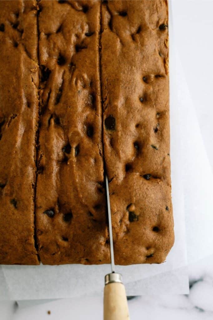 A knife is slicing through a large rectangular piece of baked chocolate chip cookie dough that is placed on a white surface.