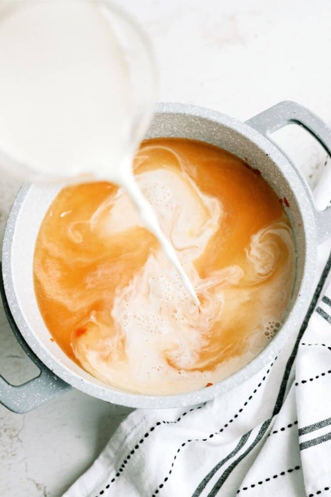 Milk being poured into a pot of tomato soup, with a striped kitchen towel beside it.