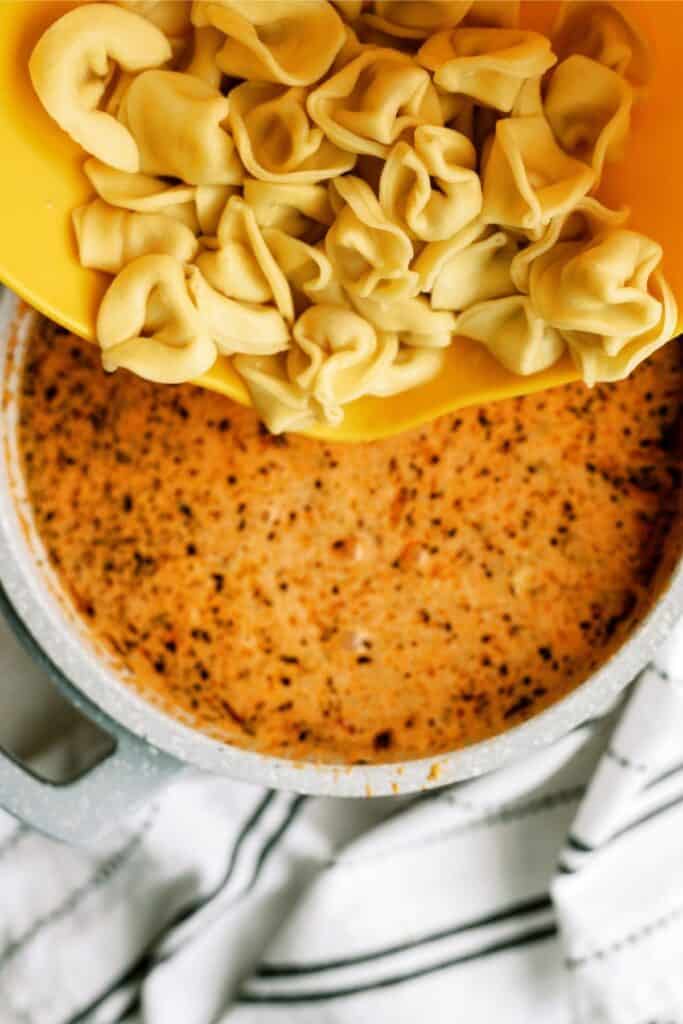 A pot of creamy, seasoned soup with tortellini being poured in from a yellow bowl above.