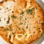 A bowl of creamy tomato tortellini soup garnished with herbs and grated cheese, served with slices of bread.