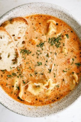 A bowl of creamy tomato tortellini soup garnished with herbs and grated cheese, served with slices of bread.