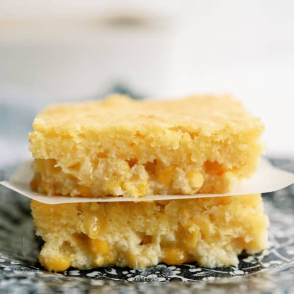 Two pieces of cornbread casserole are stacked on a plate. The cornbread has a golden yellow color and visible corn kernels.