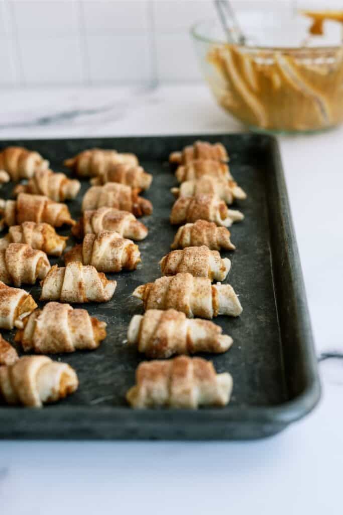 A baking tray filled with small crescent-shaped pastries sprinkled with cinnamon. A partially mixed bowl of filling sits blurred in the background.