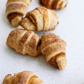 Small, golden-brown, crescent-shaped pastries sprinkled with cinnamon and sugar on a white surface.