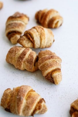 Small, golden-brown, crescent-shaped pastries sprinkled with cinnamon and sugar on a white surface.