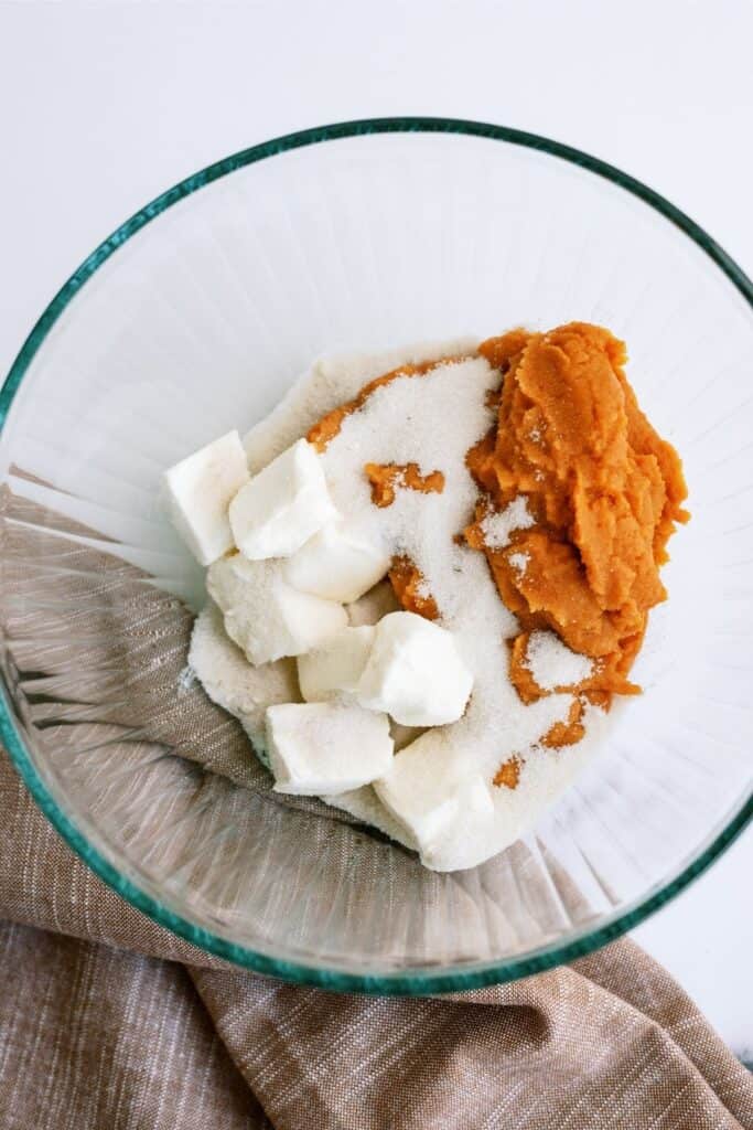 A glass bowl contains cubes of butter, granulated sugar, and a portion of pumpkin puree on a brown cloth napkin.