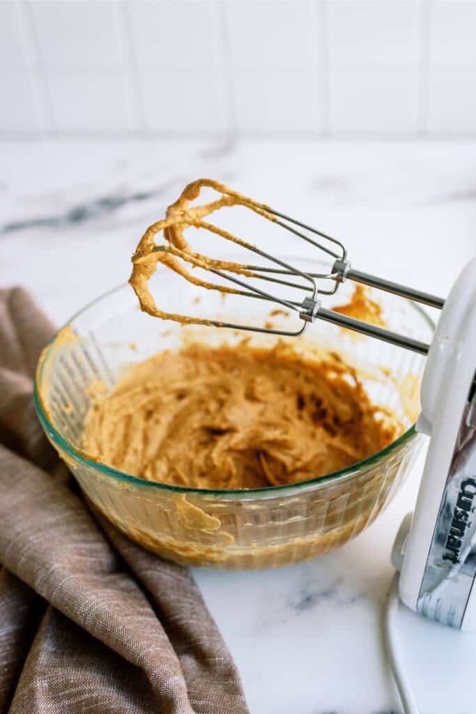 A glass mixing bowl with tan batter sits next to an electric hand mixer with batter on the beaters.