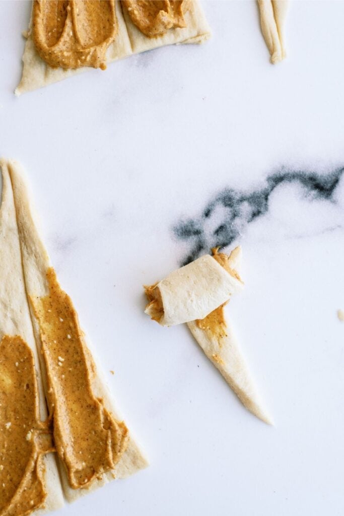 Raw dough pieces spread with peanut butter, with one piece partially rolled, placed on a white surface.