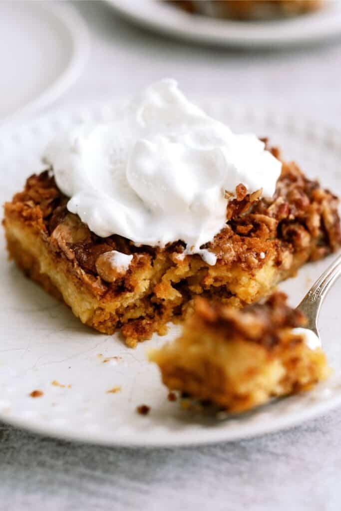 A slice of cake topped with whipped cream sits on a white plate, with a fork holding a small piece of the cake in the foreground.