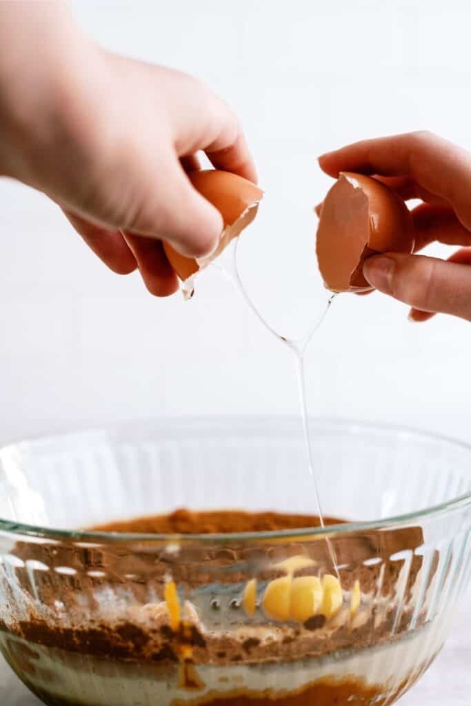 Two hands are breaking an egg over a mixing bowl containing other ingredients.