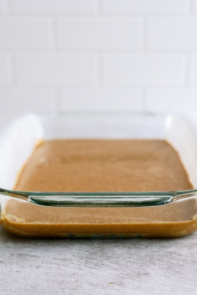 A glass baking dish with a thin layer of uncooked batter placed on a light gray countertop. The background features white subway tiles.