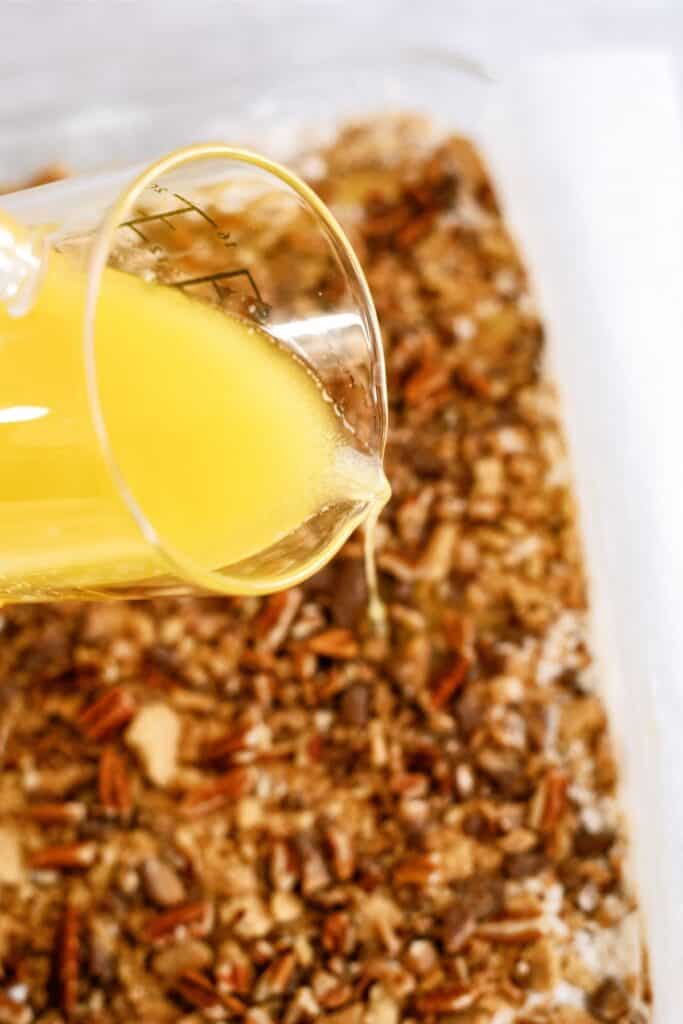 A beaker pouring melted butter over a tray of chopped pecans and other mixed ingredients.