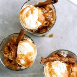 Three glass cups filled with a dessert, each topped with a scoop of vanilla ice cream, caramel sauce, and a cinnamon stick.