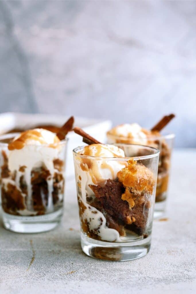 Three glass cups filled with layered dessert, featuring whipped cream, crumbled cake, caramel drizzle, and topped with cinnamon sticks on a light gray surface.