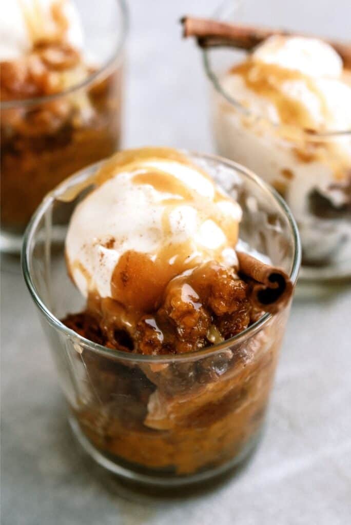 Three glass cups filled with a dessert topped with a scoop of ice cream and drizzled with caramel sauce, garnished with a cinnamon stick.