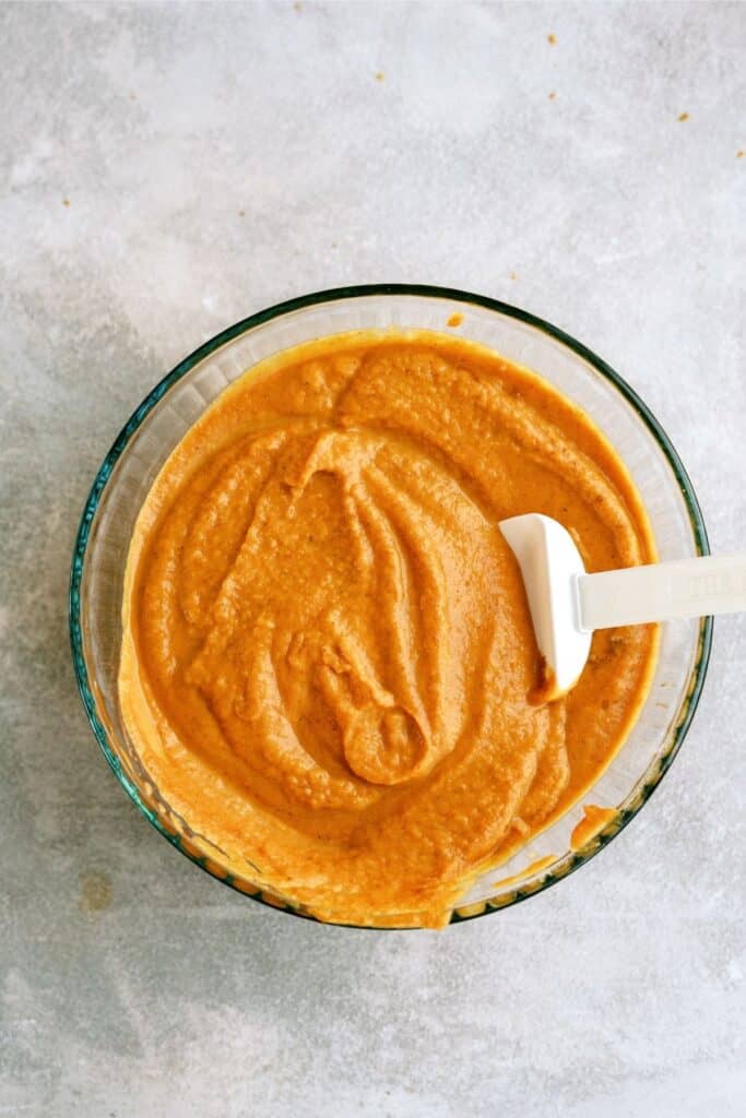 A large bowl with beaten eggs, evaporated milk, pumpkin puree, granulated sugar, brown sugar, flour, cinnamon, salt, ginger, cloves, and nutmeg mixed together with a rubber spatula