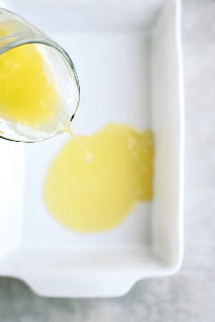 A glass pitcher is pouring yellow liquid, possibly melted butter, into a white rectangular baking dish.