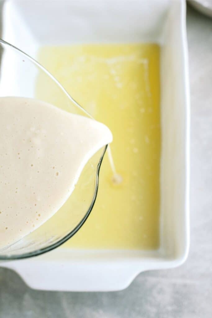 A white batter is being poured from a clear measuring cup onto a yellow layer in a rectangular white baking dish.