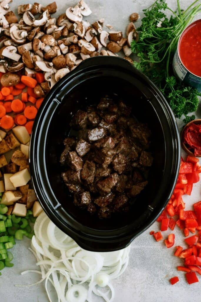 Overhead view of a slow cooker filled with cooked beef, surrounded by chopped vegetables, canned tomatoes, and herbs on a countertop.