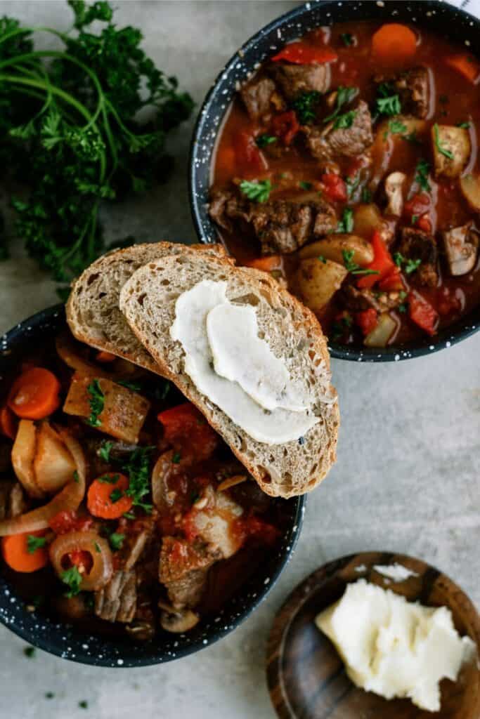 Bowls of beef stew with vegetables, garnished with parsley, a slice of bread with butter on top, and a small plate of butter on the side. Fresh parsley is in the background.