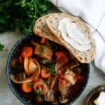 A bowl of stew with vegetables, garnished with parsley, accompanied by two slices of bread with butter. Fresh parsley and a white cloth napkin are next to the bowl.