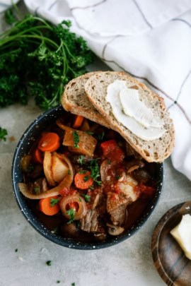 A bowl of stew with vegetables, garnished with parsley, accompanied by two slices of bread with butter. Fresh parsley and a white cloth napkin are next to the bowl.