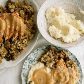 Plates of sliced turkey with gravy, mashed potatoes, and stuffing on a table, accompanied by bread rolls.