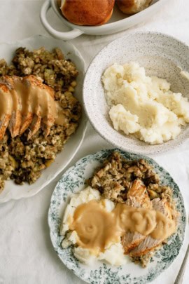 Plates of sliced turkey with gravy, mashed potatoes, and stuffing on a table, accompanied by bread rolls.