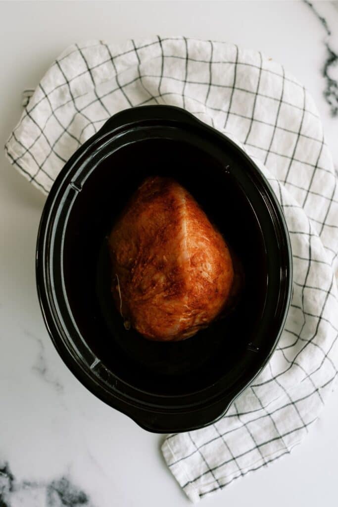 A cooked piece of meat sits in a black slow cooker, with a white checkered cloth underneath.