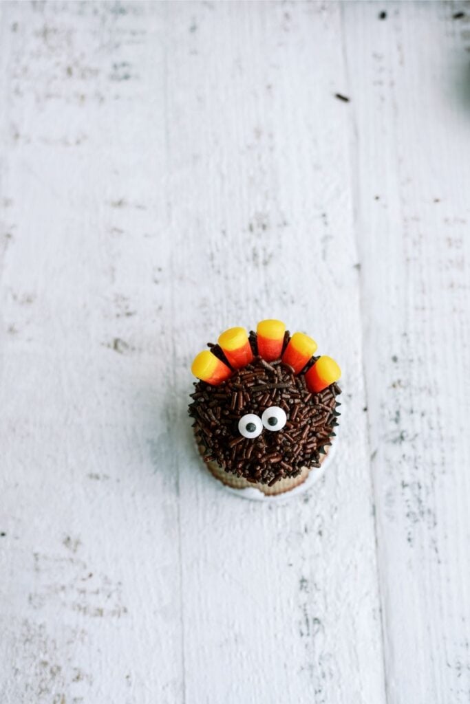 A cupcake decorated with chocolate sprinkles, candy corn, and candy eyes resembling a turkey, placed on a white wooden surface.