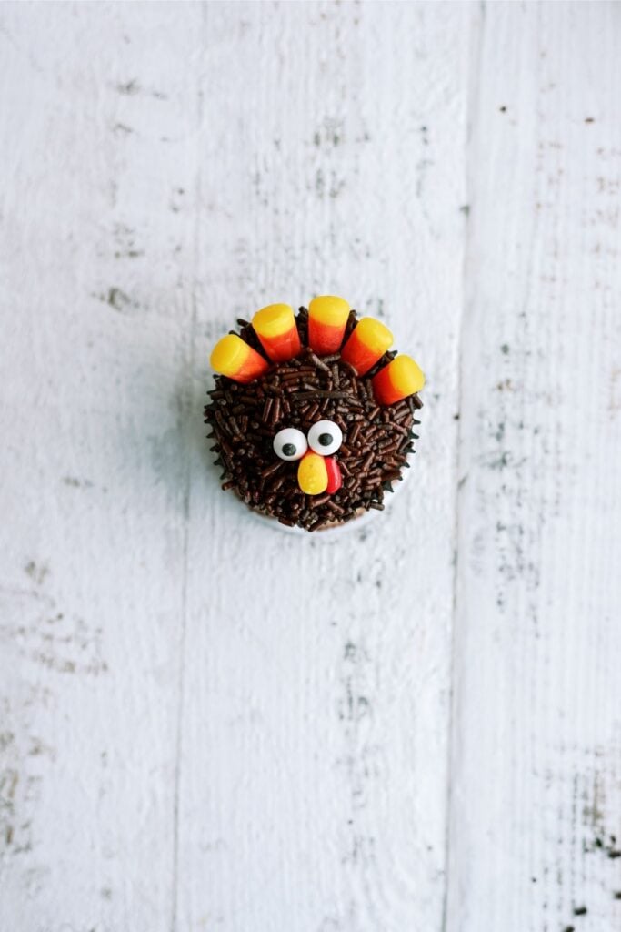 A cupcake decorated to resemble a turkey with candy eyes, candy corn feathers, and a chocolate sprinkle body on a light wooden surface.