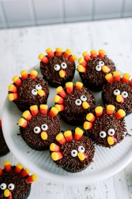 Chocolate cupcakes decorated to resemble turkeys, with candy corn feathers and candy eyes, arranged on a white plate.