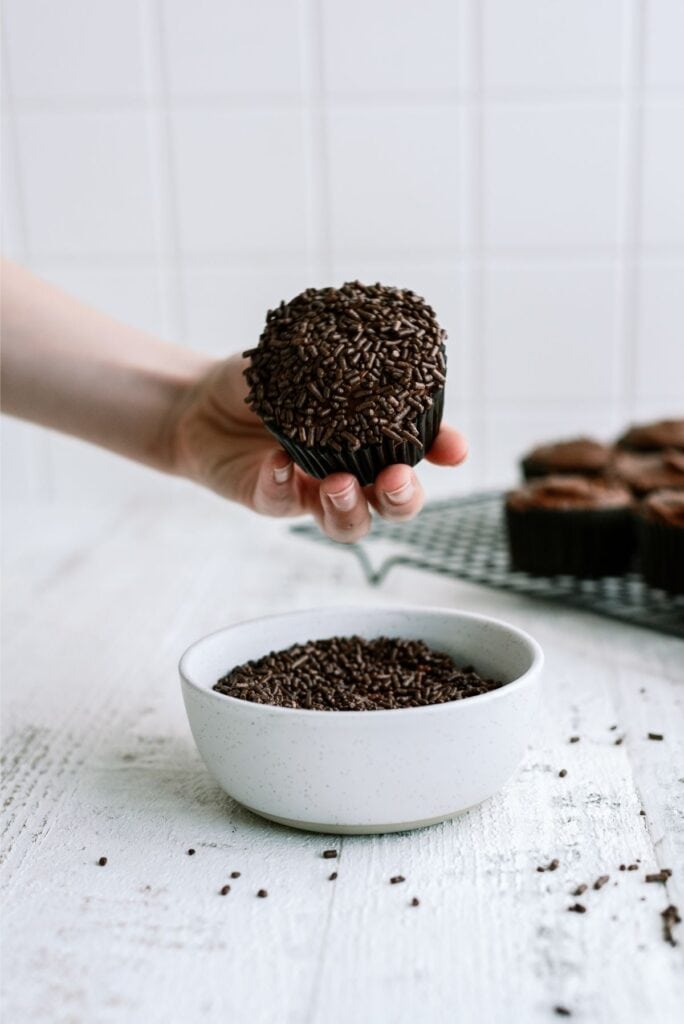 A hand holding a chocolate cupcake with sprinkles over a bowl of chocolate sprinkles. More cupcakes are on a cooling rack in the background.