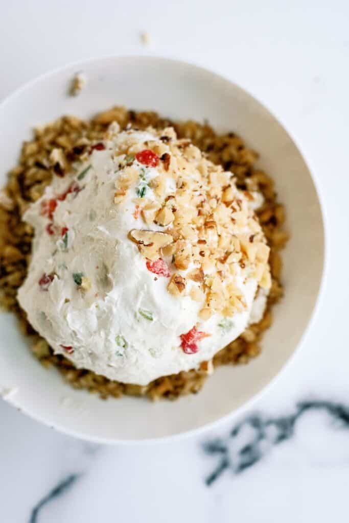 A cheese ball topped with chopped nuts and red pepper bits, served on a plate with more nuts around it.