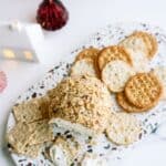 Cheese ball coated in nuts with assorted crackers on a textured platter, surrounded by small decorative houses and Christmas trees.