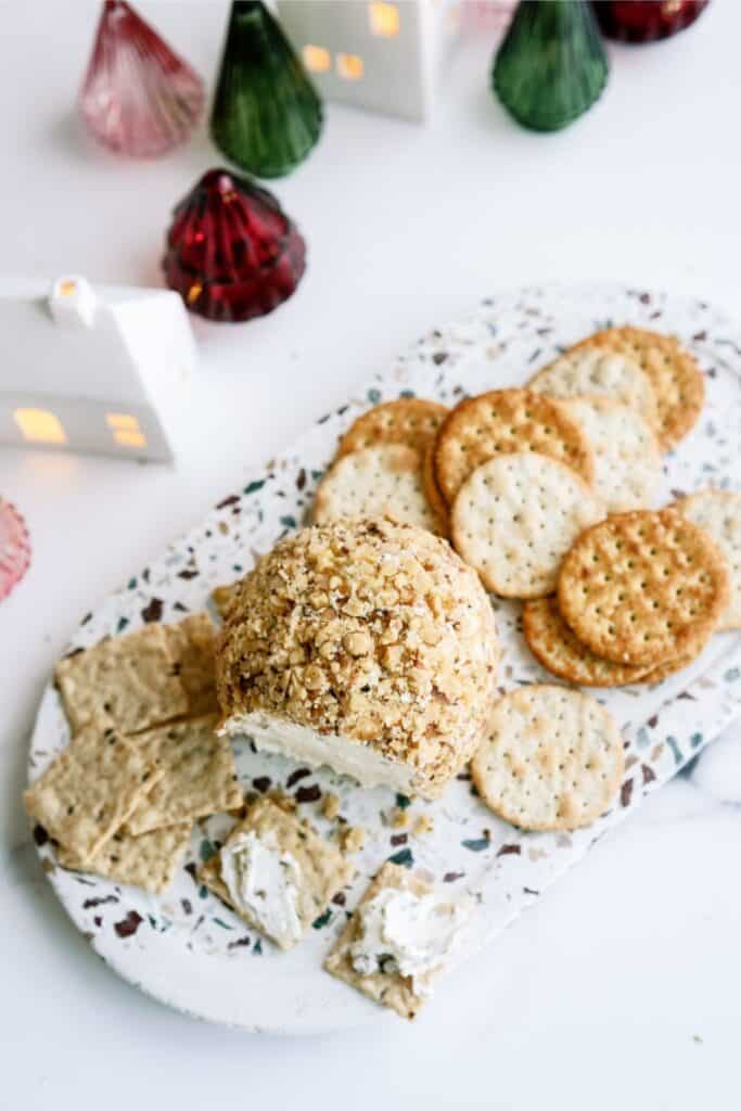 Cheese ball coated in nuts with assorted crackers on a textured platter, surrounded by small decorative houses and Christmas trees.