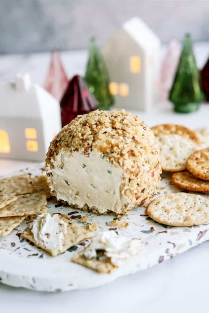 Cheese ball coated with nuts, partially eaten, on a marble board with assorted crackers. Decorative houses and trees in the background.