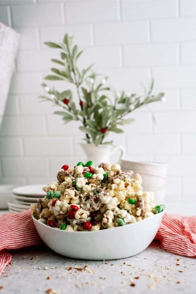 A bowl of popcorn mixed with red and green candies on a table with a red and white cloth. A small plant and stacked plates are in the background.