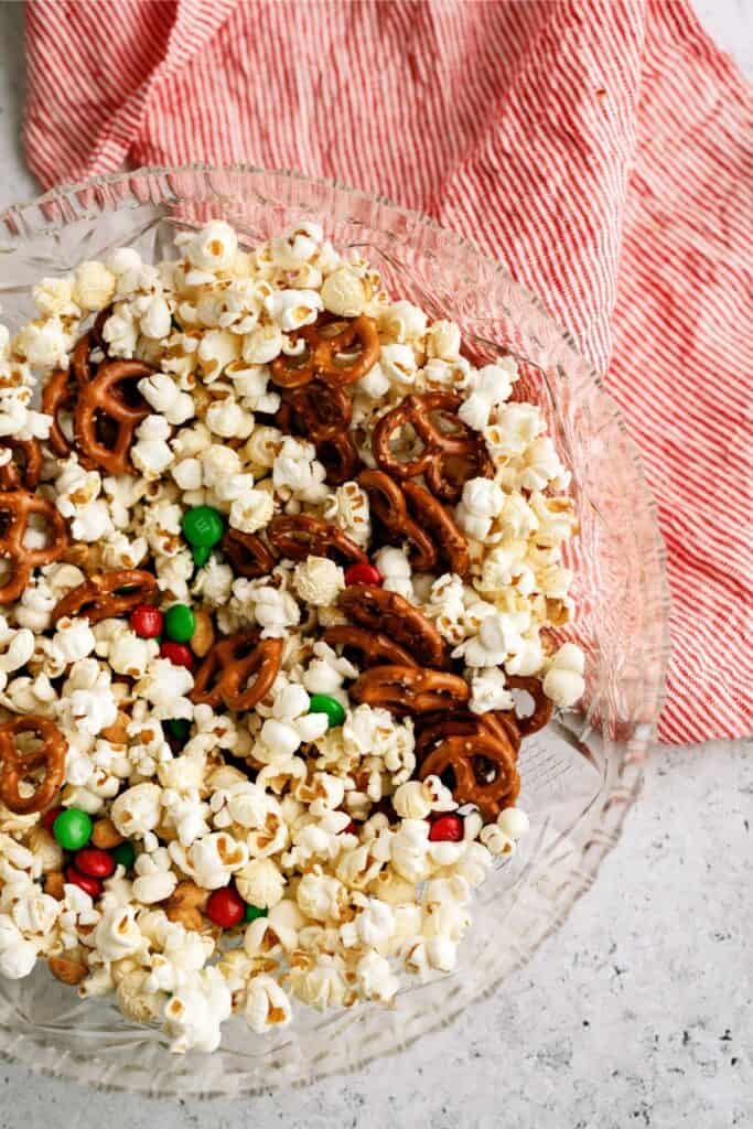 A glass bowl filled with popcorn, pretzels, and red and green candy-coated chocolates sits on a countertop next to a red and white striped cloth.