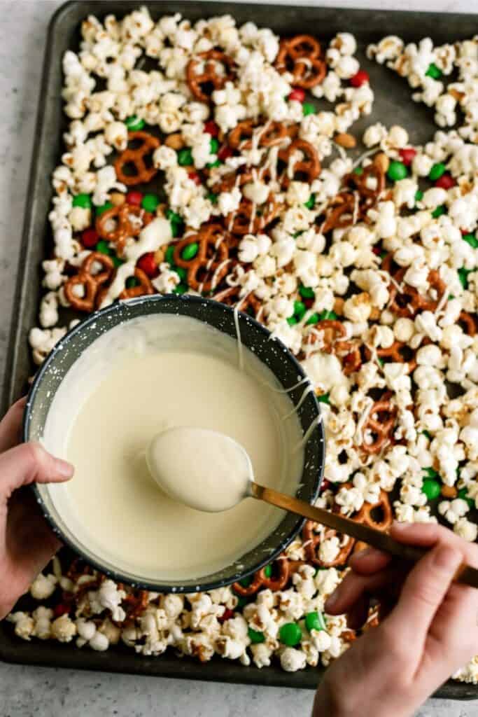 Hands holding a bowl of melted white chocolate over a baking sheet with popcorn, pretzels, and colorful candies.