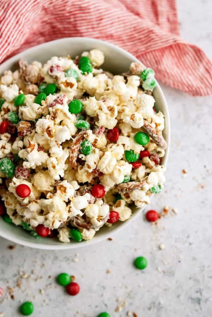 A bowl of popcorn mixed with red and green candy pieces and pretzels, placed on a light surface with a red striped cloth nearby.