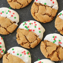 A batch of ginger cookies half-dipped in white icing, topped with green and red sprinkles, arranged on a dark surface.